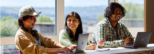 Three students sat at a desk working together with laptops